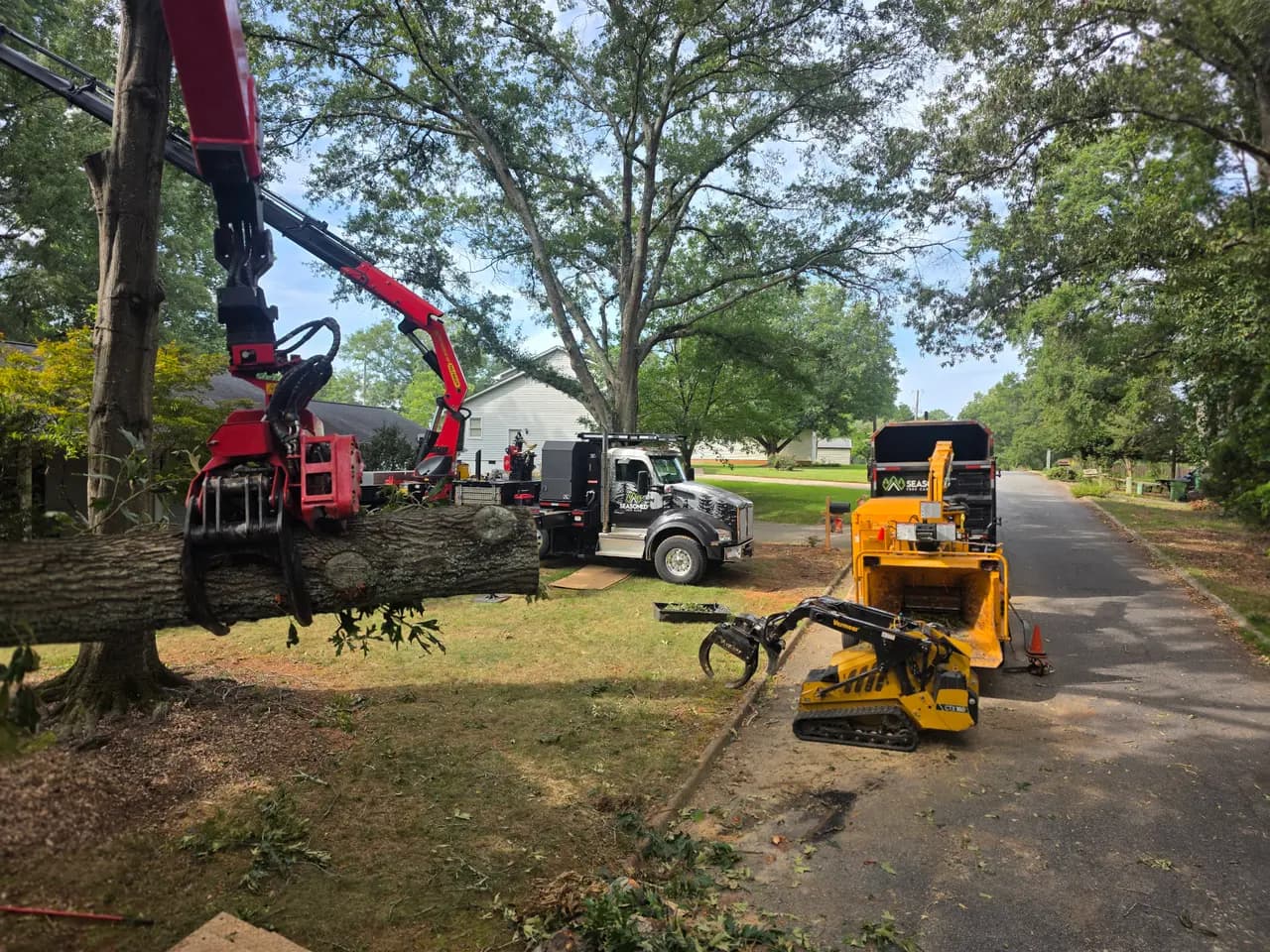 The grapple saw carrying a trunk section across the yard toward the chipper and a tracked loader.